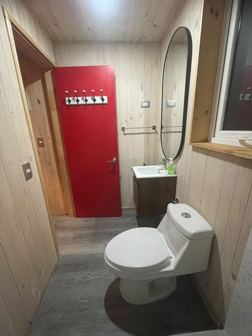 Baño de Tiny Belbén con puerta roja, espejo ovalado y piso de madera
